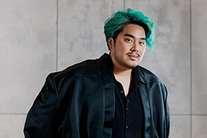 Fashion designer Bach Mai, with teal hair and a black outfit, standing in front of a light gray wall.