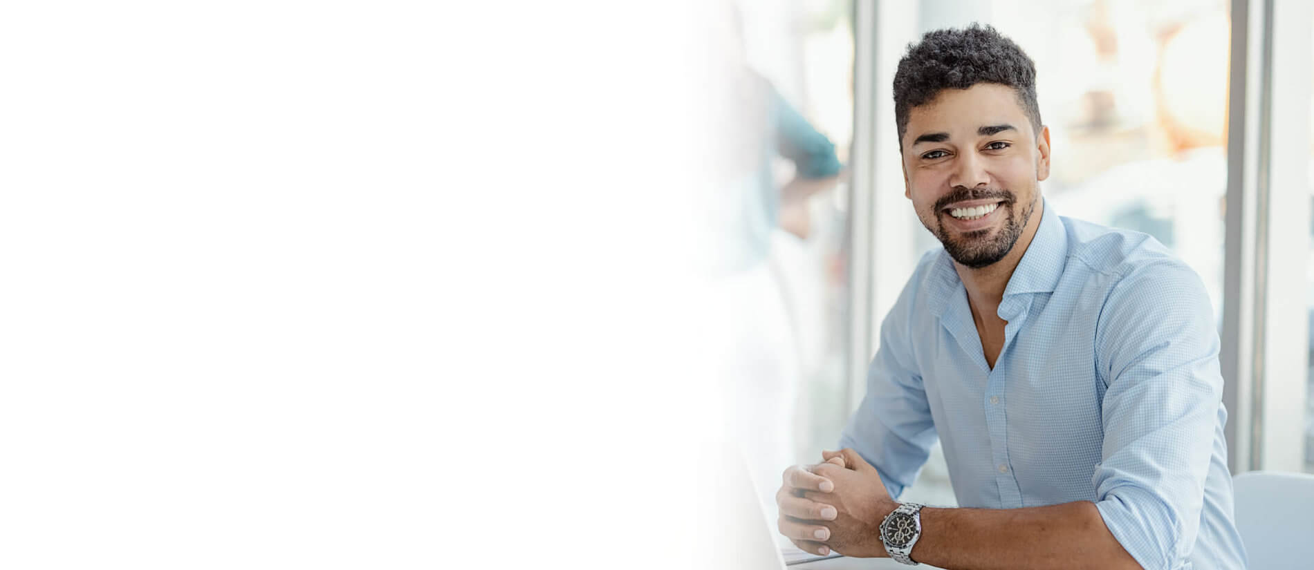 Image: A smiling young businessman in a light blue dress shirt sitting at a desk, with a white gradient fade on the left side.