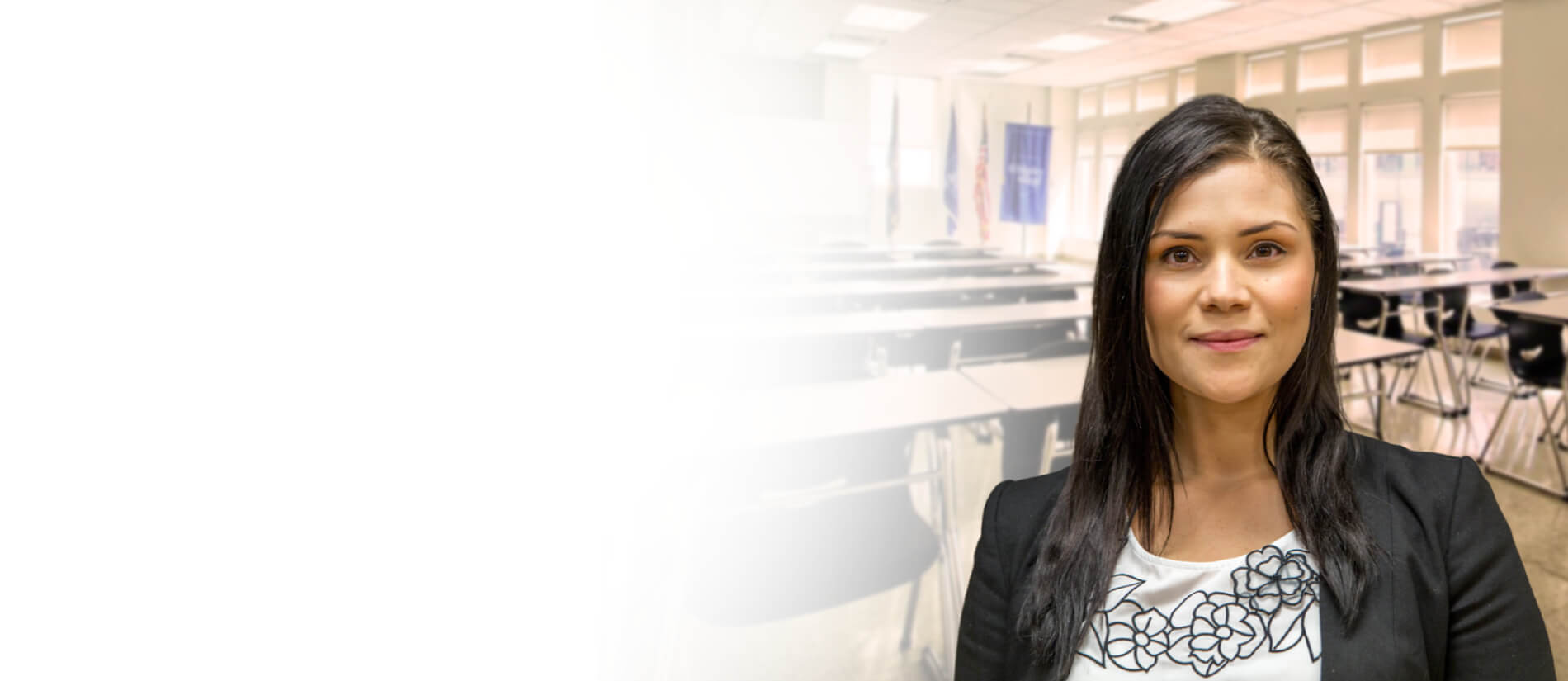 Image of Michelle Gomez standing confidently in a bright classroom, wearing a blue blazer and white shirt, conveying professionalism.