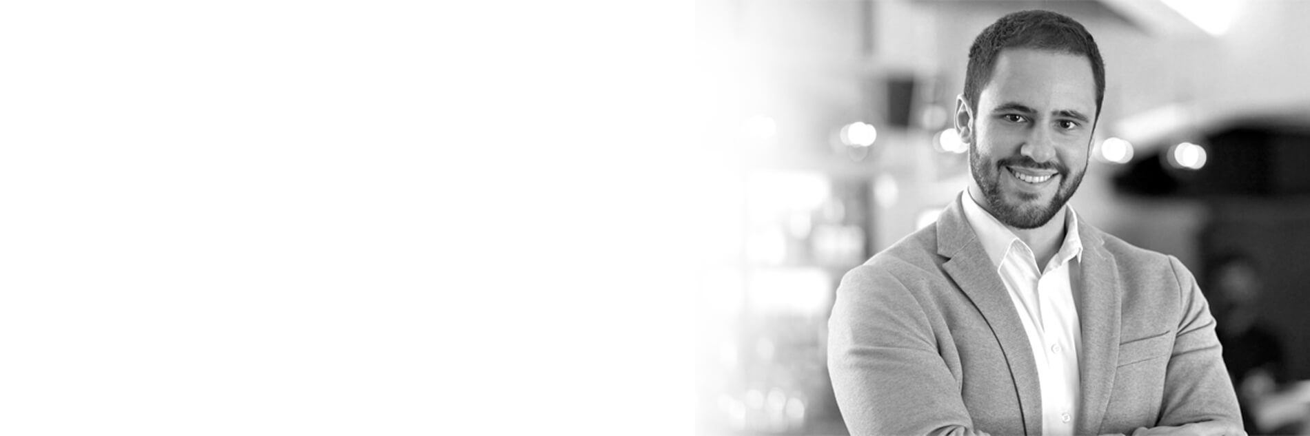 A professional man in a gray blazer stands confidently with crossed arms, set against a blurred, warm-toned background.