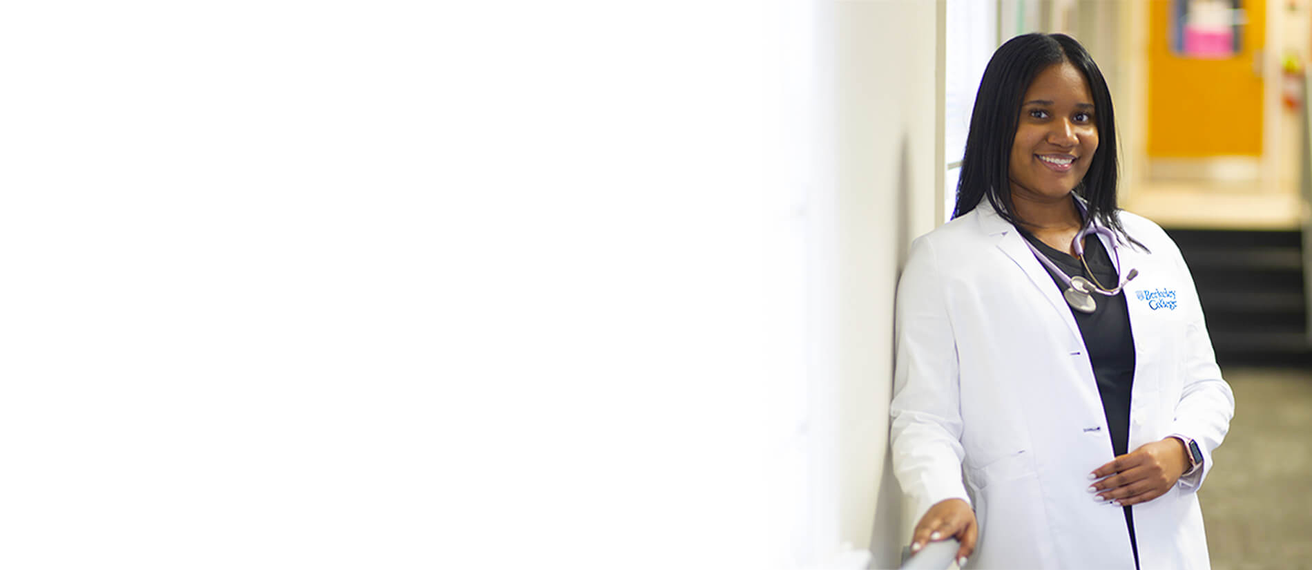A healthcare professional in a white lab coat stands confidently by a wall, wearing a stethoscope and displaying professionalism.