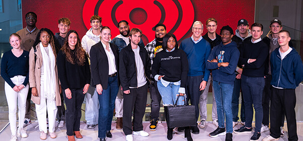 A diverse group of individuals stands together, smiling against a vibrant red iHeartRadio background. Stylish outfits showcase personal flair