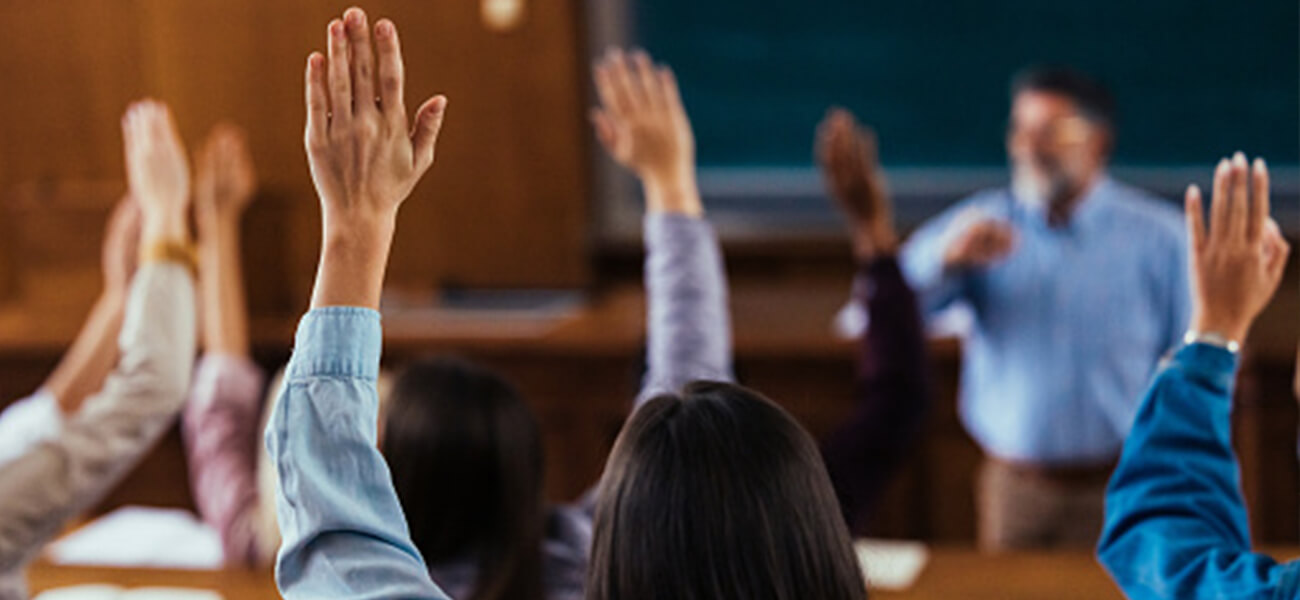 Students with raised hands are participating in a classroom discussion, focusing on the instructor at the front of the room.
