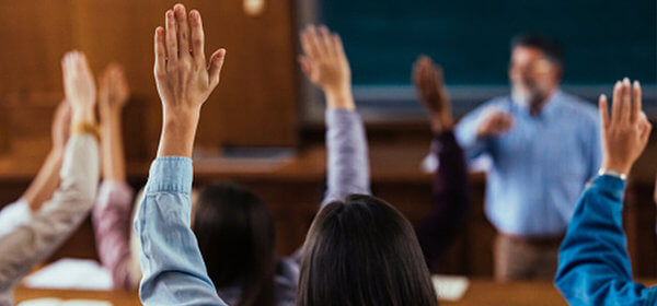 Students with raised hands are participating in a classroom discussion, focusing on the instructor at the front of the room.