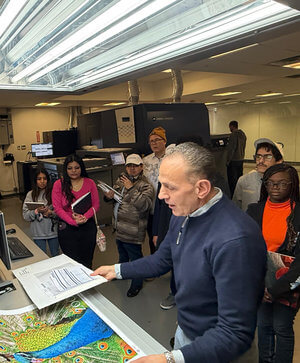 A group of people attentively listens to a man presenting printed materials in an industrial printing facility. Bright overhead lights illuminate the room.