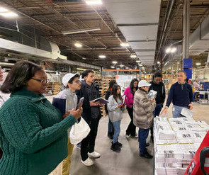 A diverse group of ten people stands smiling in a hallway with vibrant artwork on the walls. The atmosphere is friendly and welcoming.