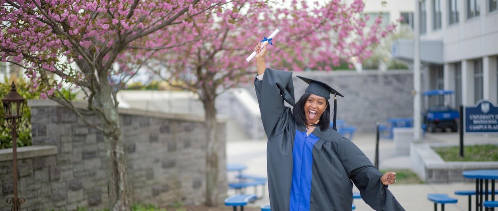 A joyful graduate in cap and gown raises her diploma triumphantly. She stands before blooming pink trees, exuding excitement and accomplishment.