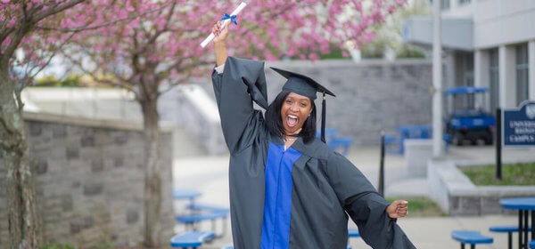 A joyful graduate in cap and gown raises her diploma triumphantly. She stands before blooming pink trees, exuding excitement and accomplishment.