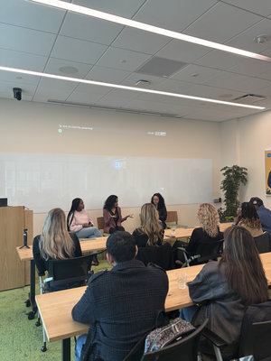 A panel discussion with three speakers on stage, addressing an audience in a modern meeting room, featuring a projector display.
