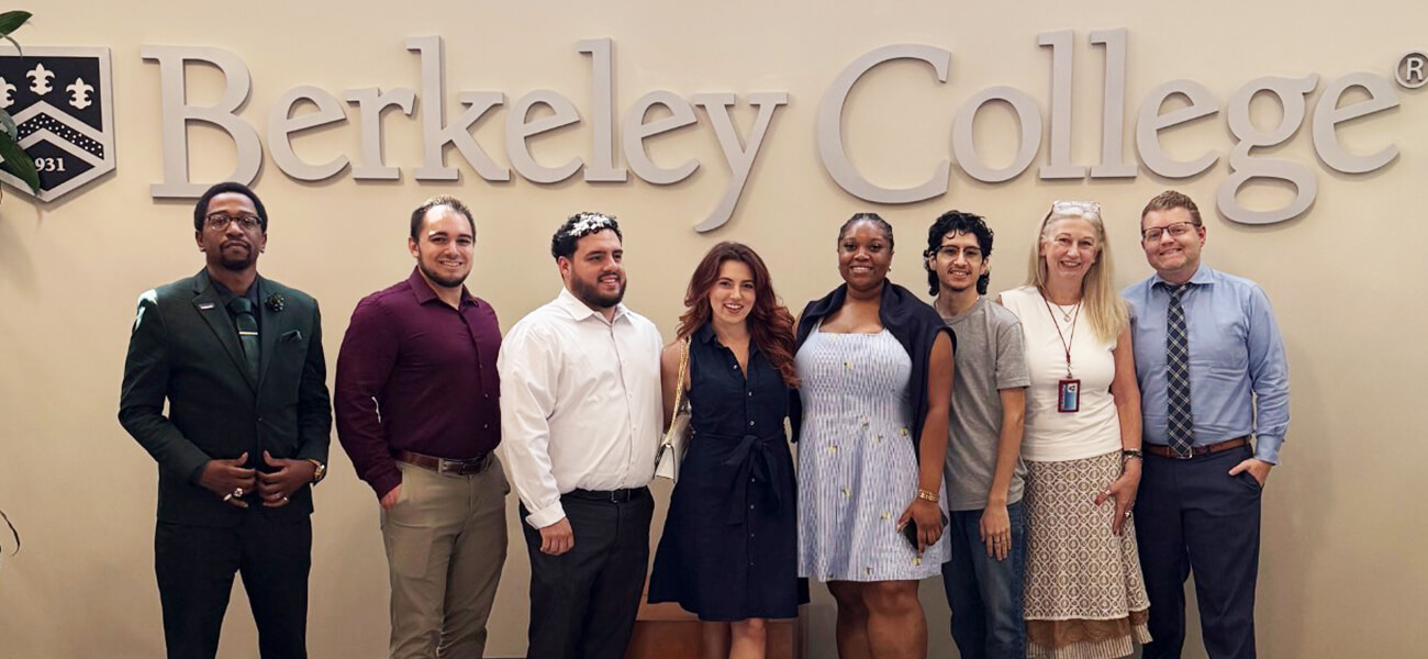 A group of eight individuals stands in front of the Berkeley College logo, smiling and dressed in various professional outfits.