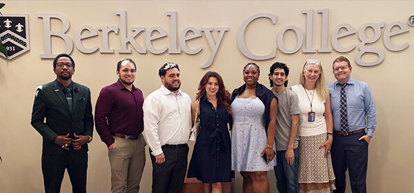 A group of eight individuals stands in front of the Berkeley College logo, smiling and dressed in various professional outfits.