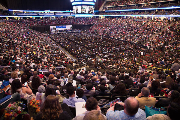 A vast arena filled with a seated crowd. The center features a stage with graduates in caps and gowns. The atmosphere is celebratory and bustling.