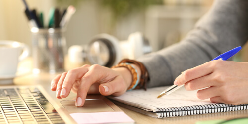 A person writes in a notebook while typing on a laptop. The space is cozy, with blurred stationery in the background, conveying focus and productivity.