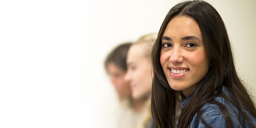 A woman is seated in a classroom, smiling brightly, reflecting engagement and enthusiasm for the learning experience