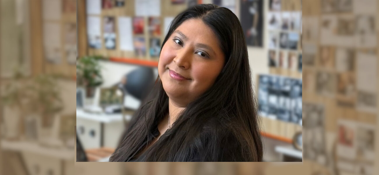 Smiling woman with long dark hair in a cozy office, featuring blurred photos and plants in the background, conveying a warm, welcoming atmosphere