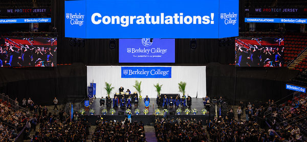 Graduation ceremony at Berkeley College with graduates on stage and a large blue: Congratulations, banner overhead. Audience seated, celebrating.