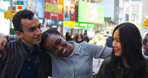 Three smiling individuals stand together on a bustling city street, surrounded by urban scenery