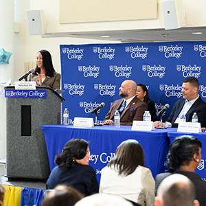 A woman stands at a podium in front of a crowd giving a speech. A panel of people sits next to her on a stage.