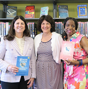 Three diverse women, two holding books, stand in a row in front of a bookshelf smiling