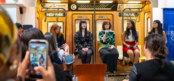 Five women sit on an orange Coach subway-car set as an audience watches and takes photos at a lively New York fashion event