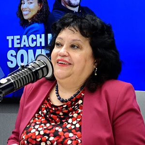 A woman in a bright floral blouse and pink blazer sits beside a microphone, with a blue background