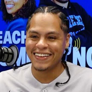 Smiling person with braided hair wearing a gray shirt, seated in front of a microphone against a blue background with partial visible text.