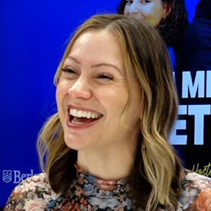 A woman with wavy hair smiles widely, wearing a floral top against a vibrant blue background with partially visible text. The mood is cheerful and upbeat.