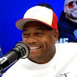 A person wearing a red and white cap is seated in front of a microphone, with a blue background behind them.