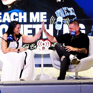 A woman in a white dress and a man in a black shirt high-five each other during a live talk show on stage with a colorful backdrop.