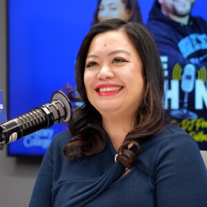 Photo of Lourdes Nanong, Smiling woman with long dark hair sits in a recording studio, wearing a blue top. A microphone is in front of her, and a colorful backdrop displays behind.