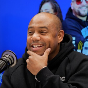 Smiling man in a black hoodie sits at a microphone with a thoughtful expression. A vibrant blue background features blurred people, adding a lively mood.