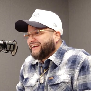 A person wearing a plaid shirt and a cap sits in front of a microphone, engaged in a recording or discussion.