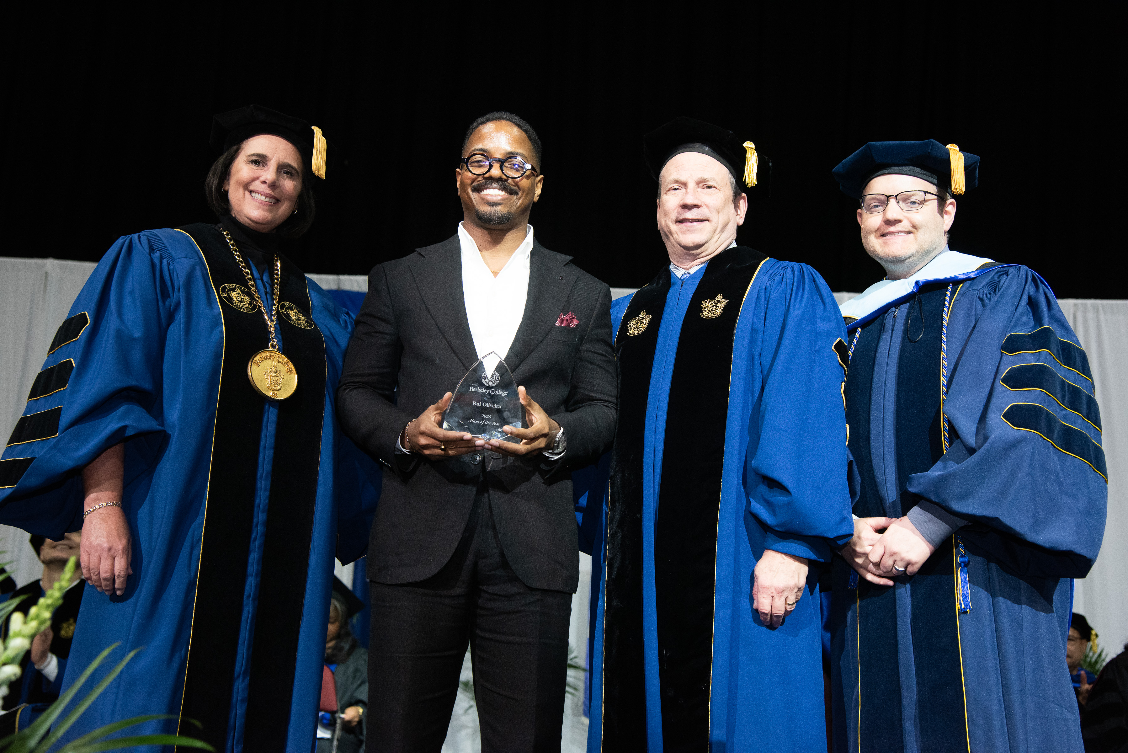 Berkeley College officials stand with a man who is Alumnus of the Year, in regalia on stage.
