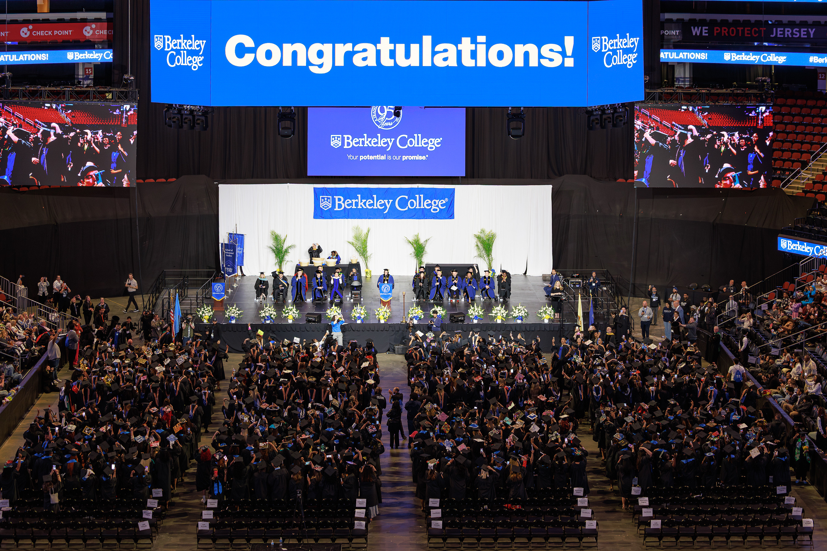 Graduation ceremony at Berkeley College with graduates in caps and gowns, faculty on stage, and a large "Congratulations!" sign overhead.