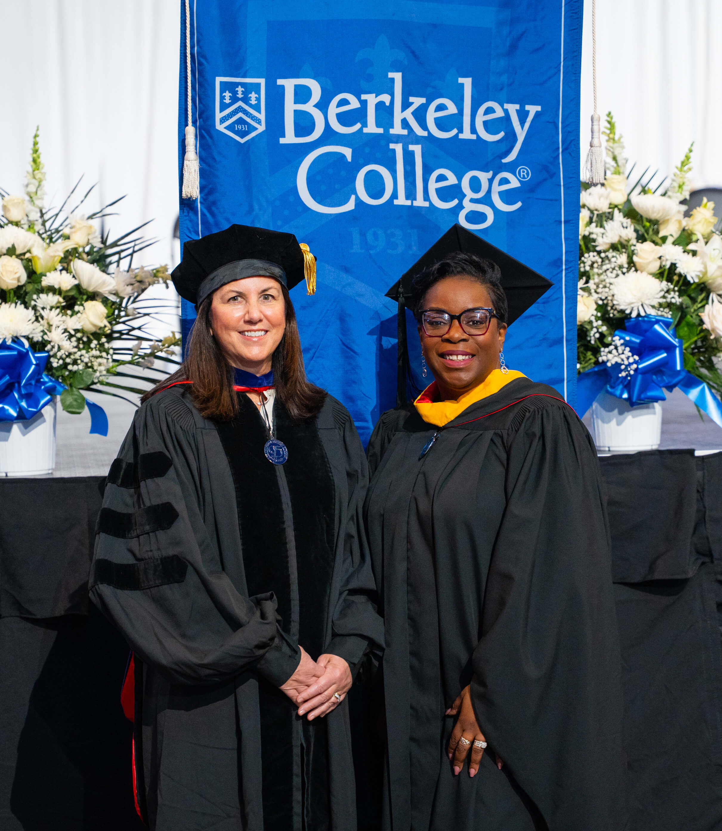 Two women in regalia pose in front of a Berkeley College blue banner.