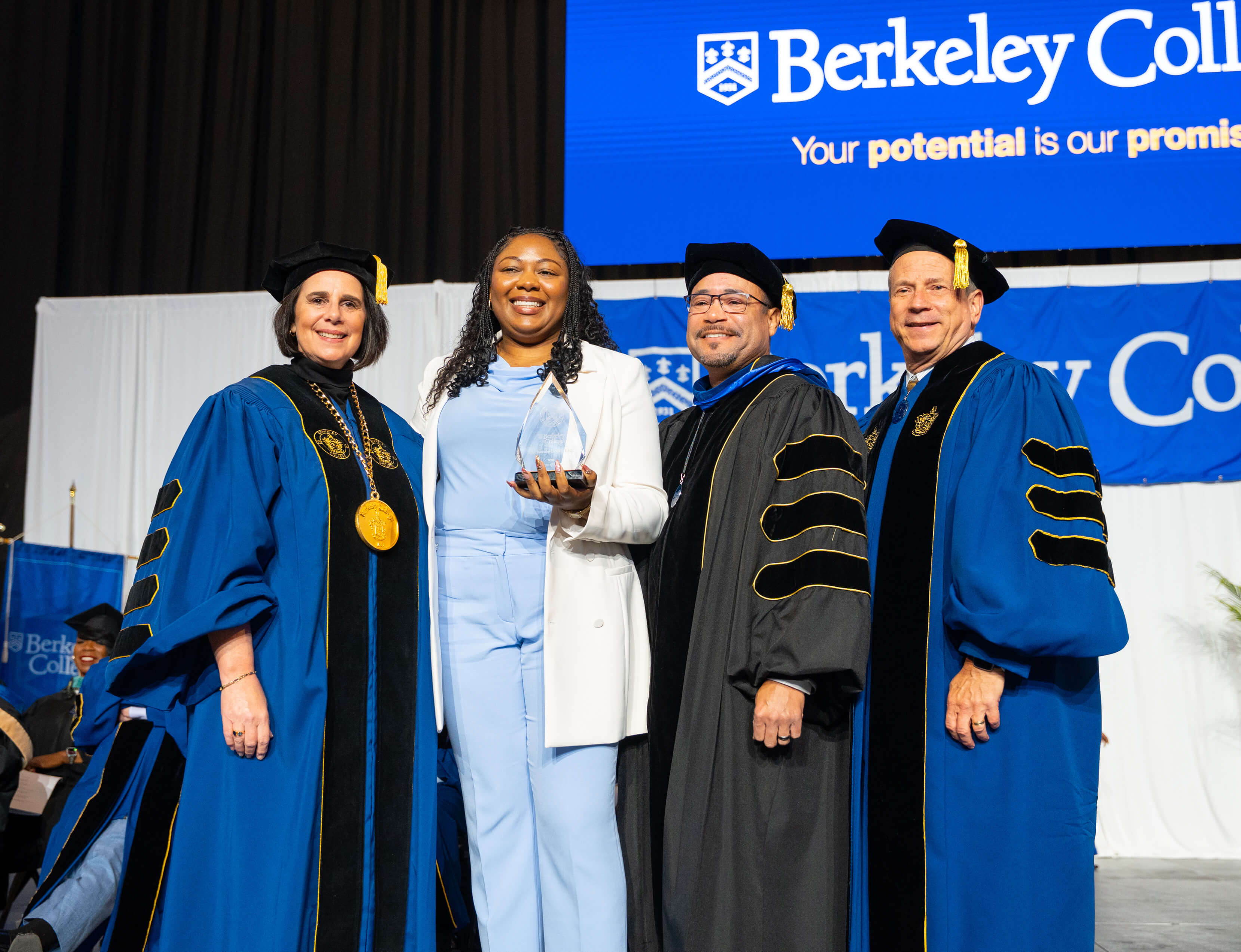 Four people standing on stage at a Berkeley College event, with one person holding an award and others in academic regalia.