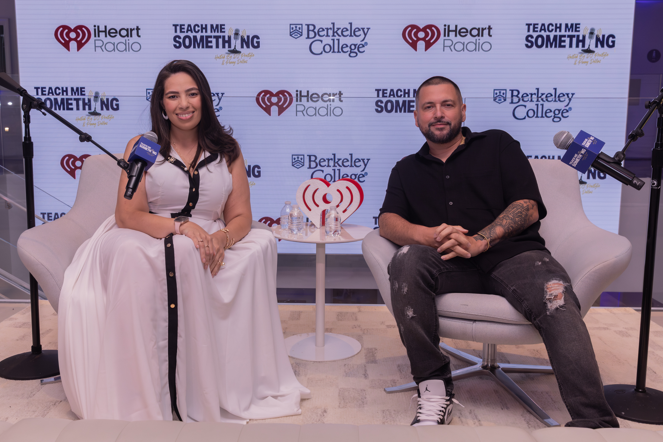A woman and a man sit in front of a backdrop with microphones