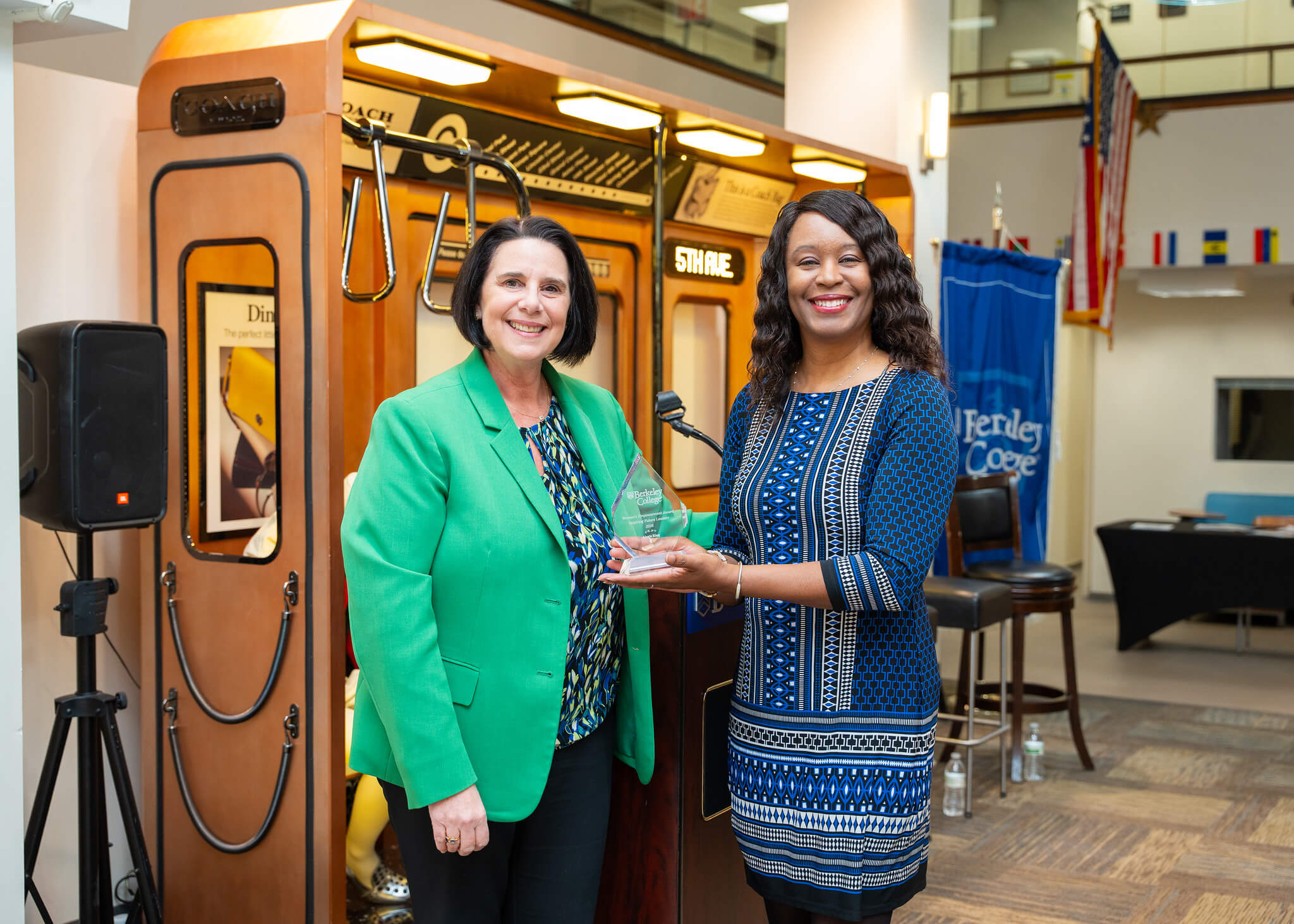Two smiling women pose with a Berkeley College award in front of a podium at an indoor event.