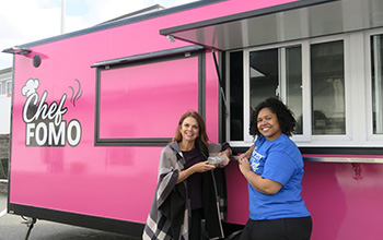 Two women stand outside of a bright pink food truck titled Chef FOMO.