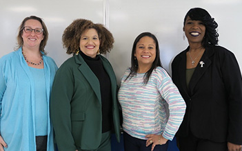 Four diverse women stand smiling in front of a whiteboard.