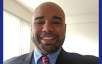 Smiling man with a trimmed beard wearing a dark suit, blue shirt, and red striped tie in a bright room with vertical blinds.