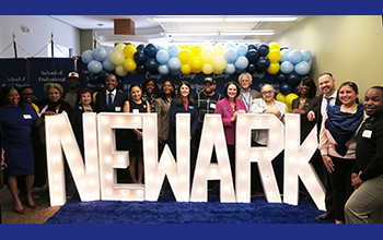 Group of people posing behind large illuminated letters spelling "NEWARK" with blue, white, and yellow balloons in the background.