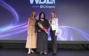 Three women pose onstage at a WBLF event; the center woman holds a bouquet beneath WBLF and NJBIA signage.