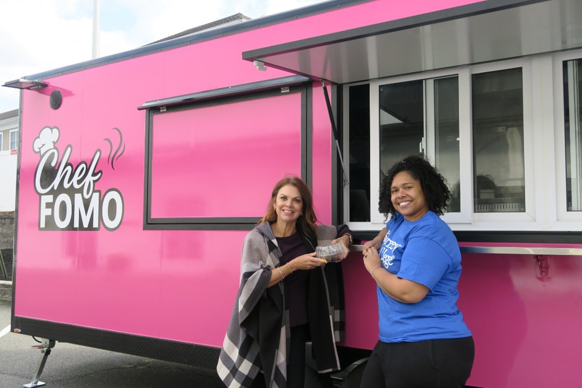 Two women stand outside of a bright pink food truck labeled CHEF FOMO.