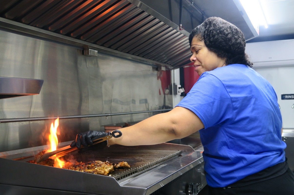 A woman stands over a flaming grill cooking chicken