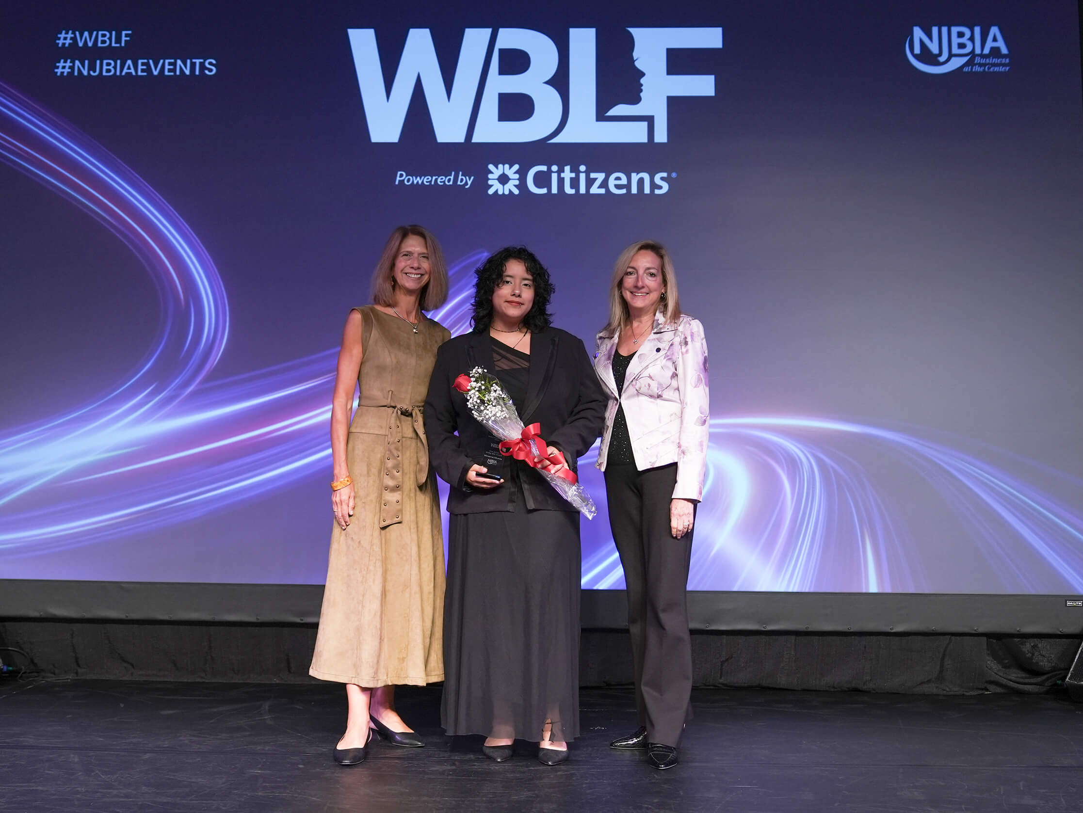 Three women pose onstage at a WBLF event; the center woman holds a bouquet beneath WBLF and NJBIA signage.