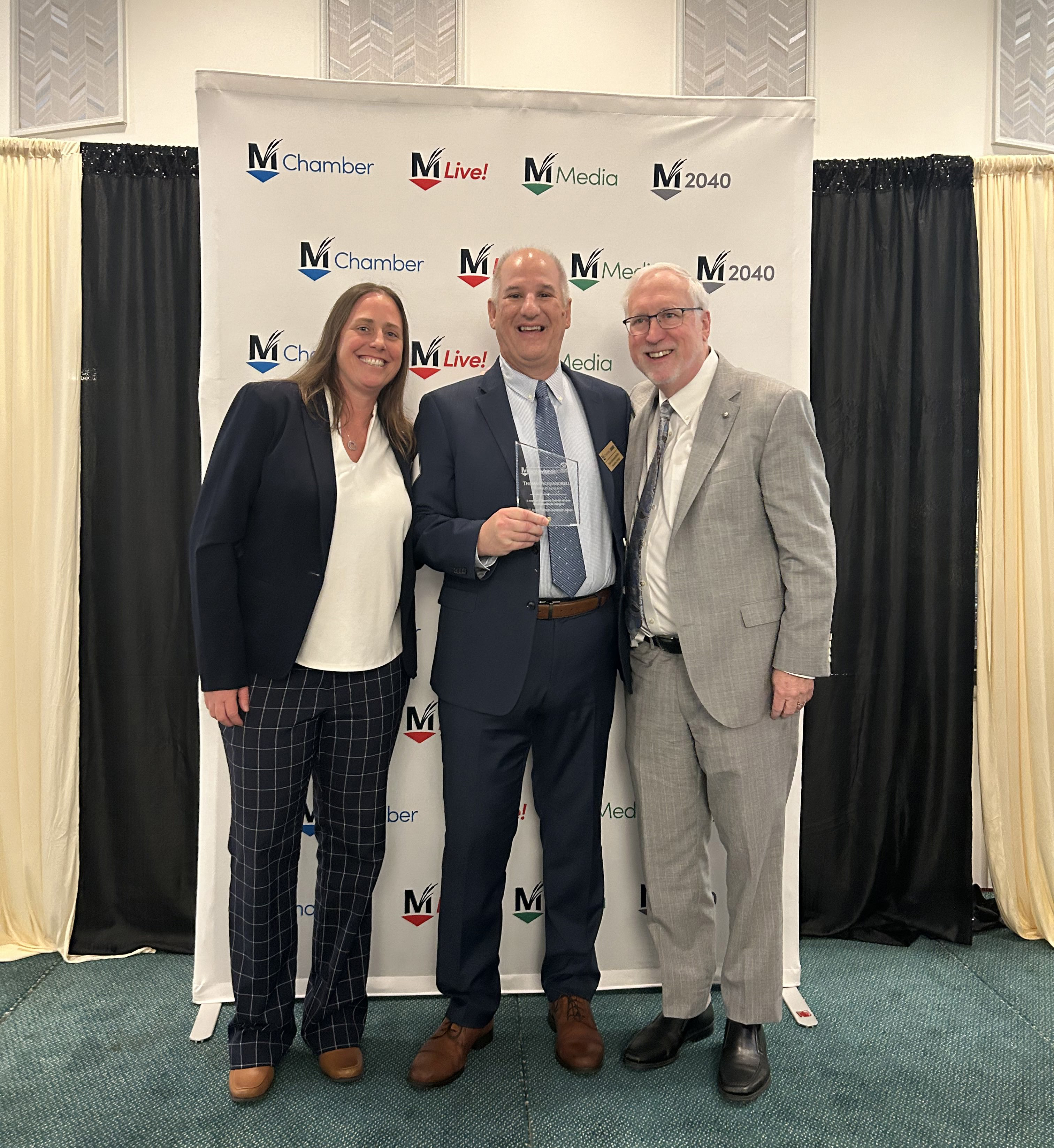 Three individuals stand in front of a chamber of commerce backdrop.