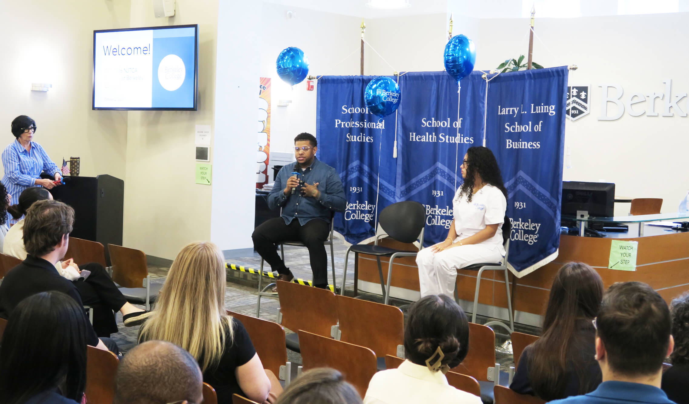 Panel discussion at Berkeley College, with two speakers seated before branded banners and an audience watching.