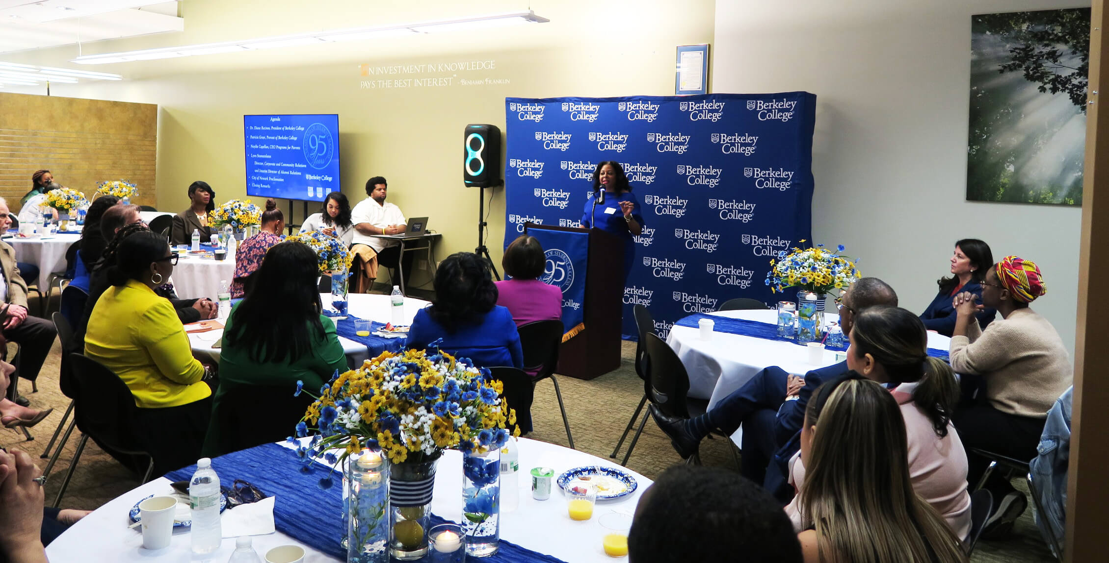 Audience attentively listens to a speaker at a Berkeley College event celebrating 95 years, with floral centerpieces and a presentation screen.