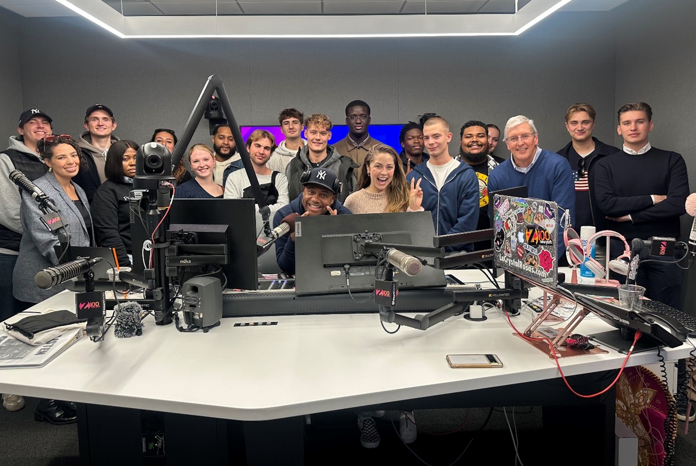Students, associates, and radio hosts gather around a desk and microphone